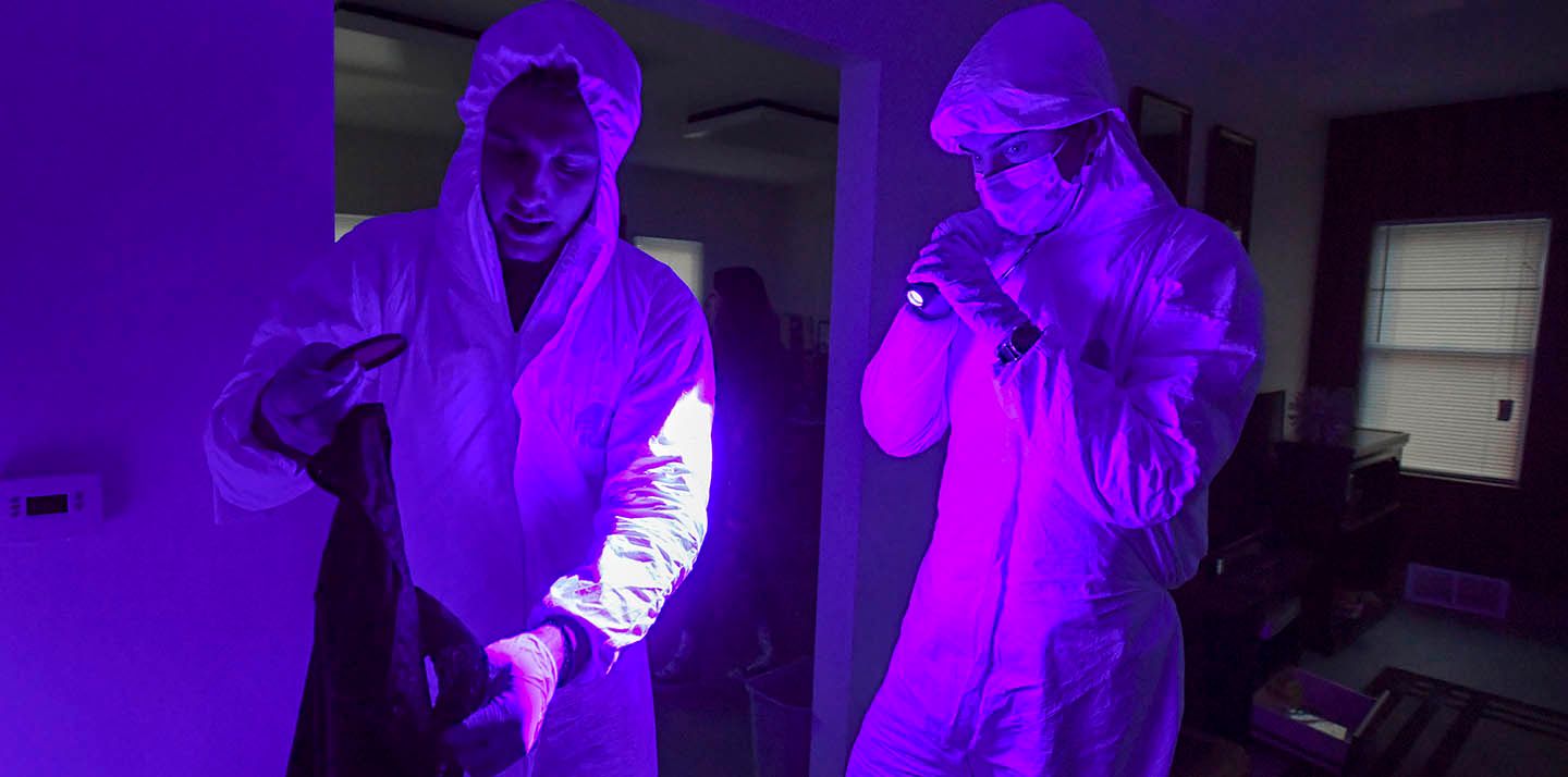 Two people in protective gear using a blacklight on a scene in a dark house