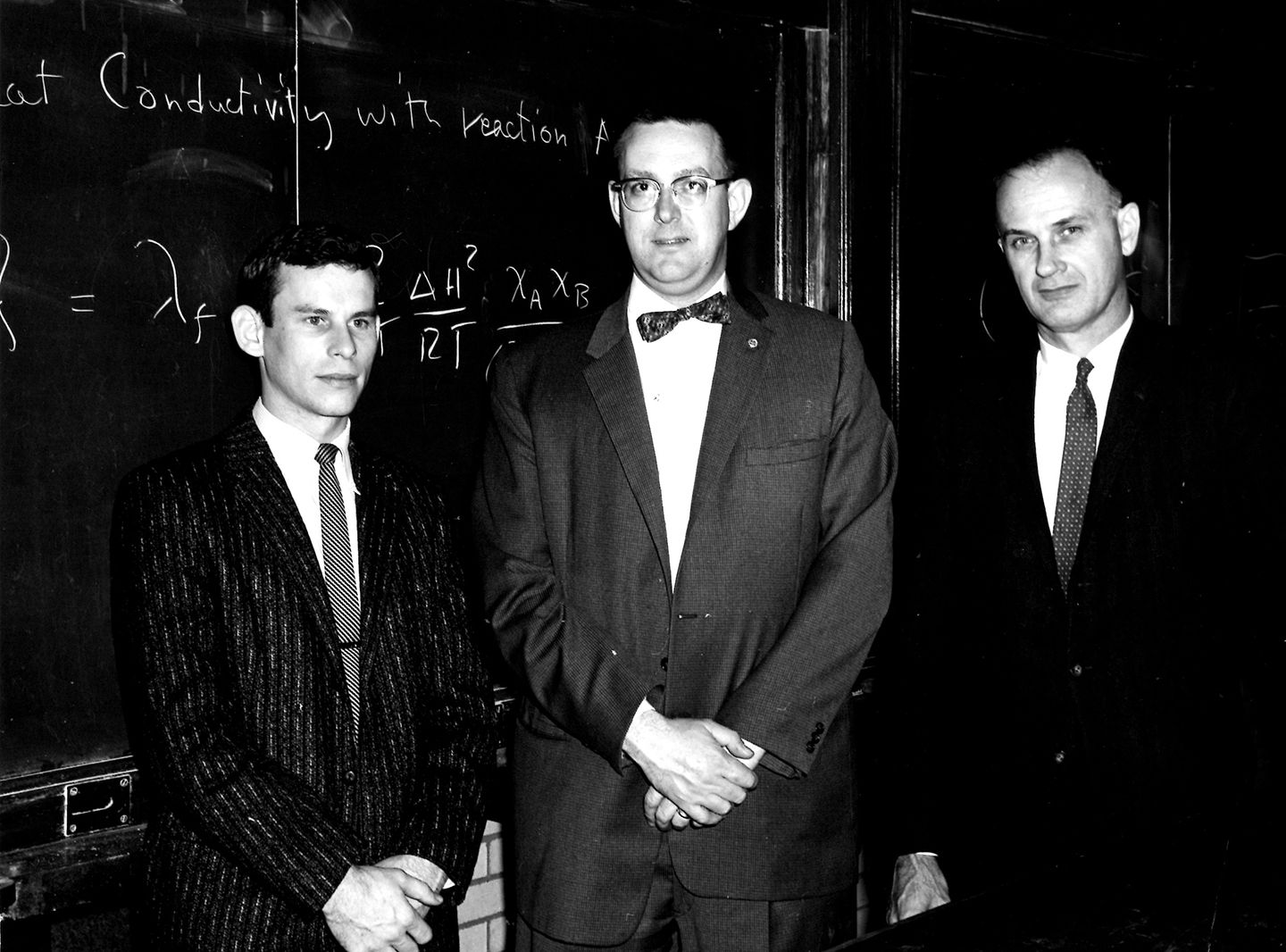 Three men in an old photo standing in front of a chalkboard wearing suits